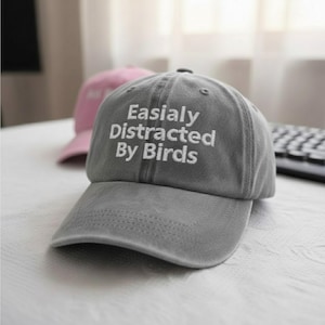 May include: Grey baseball cap with the embroidered text "Easialy Distracted By Birds" in white. A pink baseball cap is visible in the background. The cap has a curved peak and a structured crown.