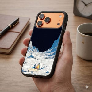 Puede incluir: Una funda de smartphone negra con una ilustración gráfica de un paisaje lunar con una pequeña tienda de campaña y un astronauta. La parte superior de la funda tiene una carcasa de cámara naranja metálica con tres lentes circulares.