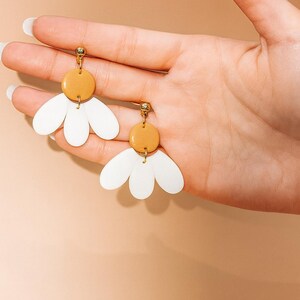Puede incluir: Un par de pendientes con forma de margarita. Cada pendiente presenta una parte superior circular de color mostaza y tres elementos blancos en forma de p&eacute;talo. Los pendientes se sostienen en una mano sobre un fondo color melocot&oacute;n.