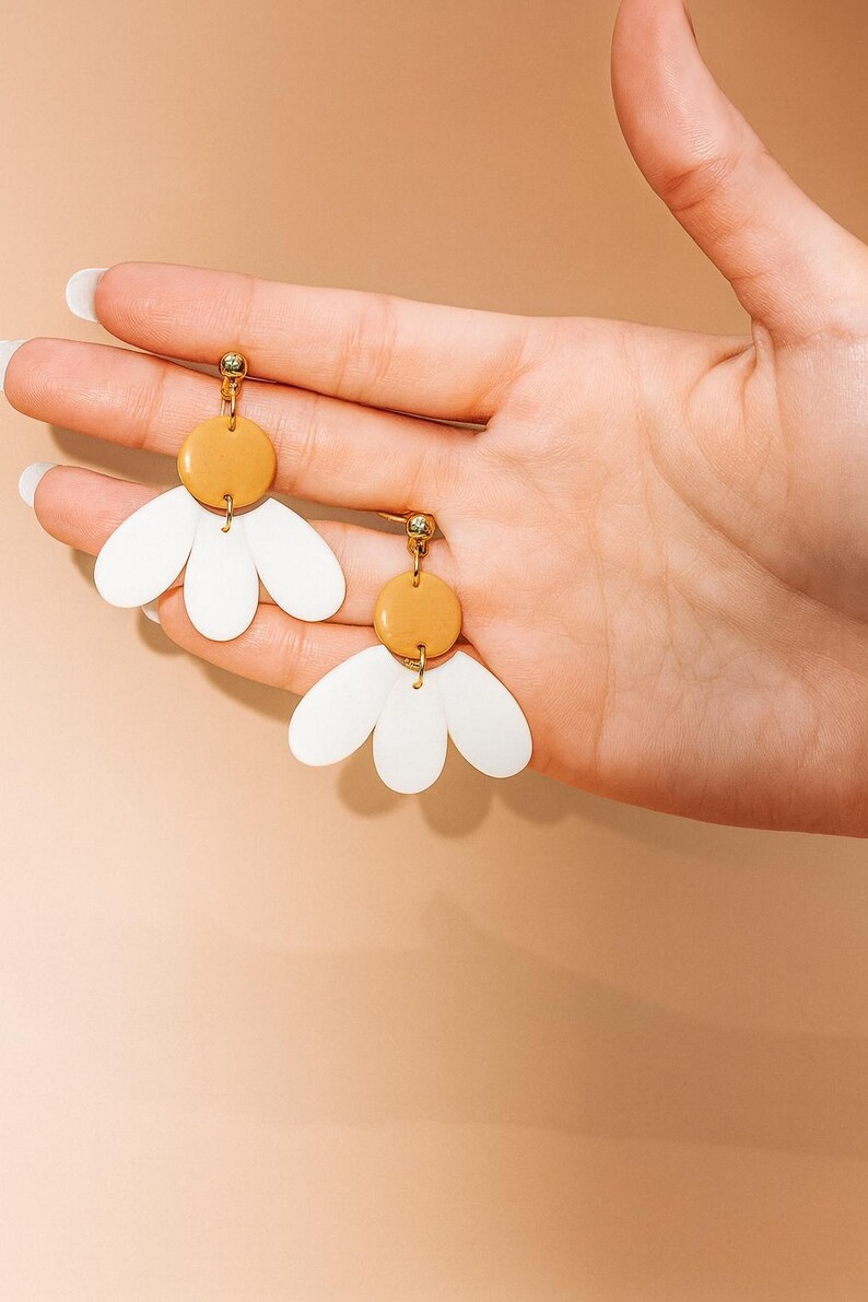 Puede incluir: Un par de pendientes con forma de margarita. Cada pendiente presenta una parte superior circular de color mostaza y tres elementos blancos en forma de p&eacute;talo. Los pendientes se sostienen en una mano sobre un fondo color melocot&oacute;n.