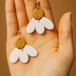 Puede incluir: Un par de pendientes hechos a mano con un perno dorado, un disco color mostaza y tres elementos blancos en forma de p&eacute;talo. Los pendientes se sostienen en una mano sobre un fondo c&aacute;lido y neutro. Dise&ntilde;o floral minimalista.