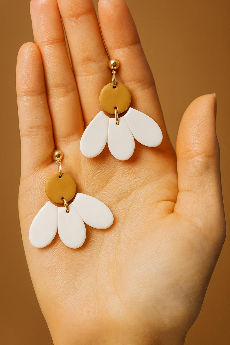 Puede incluir: Un par de pendientes hechos a mano con un perno dorado, un disco color mostaza y tres elementos blancos en forma de p&eacute;talo. Los pendientes se sostienen en una mano sobre un fondo c&aacute;lido y neutro. Dise&ntilde;o floral minimalista.