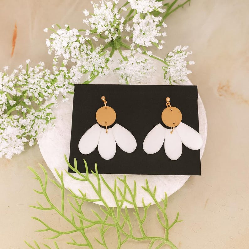 Puede incluir: Un par de pendientes blancos con forma de margarita con la parte superior circular dorada. Los pendientes se muestran sobre una tarjeta negra, con flores blancas y follaje verde en el fondo. Dise&ntilde;o sencillo y elegante.