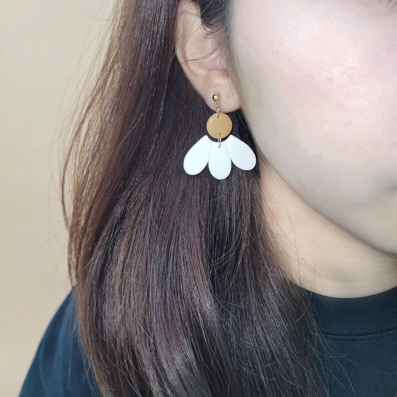 Puede incluir: Pendientes de bot&oacute;n dorados con un disco marr&oacute;n y detalles blancos en forma de p&eacute;talo. Los pendientes est&aacute;n dise&ntilde;ados para parecerse a una flor. Los pendientes se llevan en la oreja de una persona.