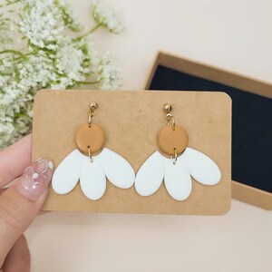 Puede incluir: Un par de pendientes hechos a mano con un dise&ntilde;o de flor blanca y un centro marr&oacute;n, con herrajes dorados. Los pendientes se exhiben en una tarjeta marr&oacute;n, con una caja de regalo en el fondo. Un accesorio &uacute;nico.