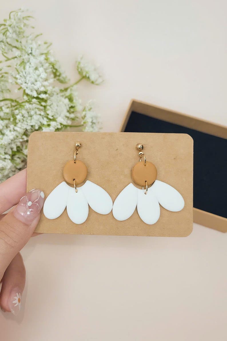 Puede incluir: Un par de pendientes hechos a mano con un dise&ntilde;o de flor blanca y un centro marr&oacute;n, con herrajes dorados. Los pendientes se exhiben en una tarjeta marr&oacute;n, con una caja de regalo en el fondo. Un accesorio &uacute;nico.