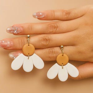 Puede incluir: Un par de pendientes dorados con una parte superior circular de color marr&oacute;n claro y una parte inferior blanca en forma de p&eacute;talo. Los pendientes est&aacute;n dise&ntilde;ados para parecerse a flores. Los pendientes se muestran sobre un fondo neutro.