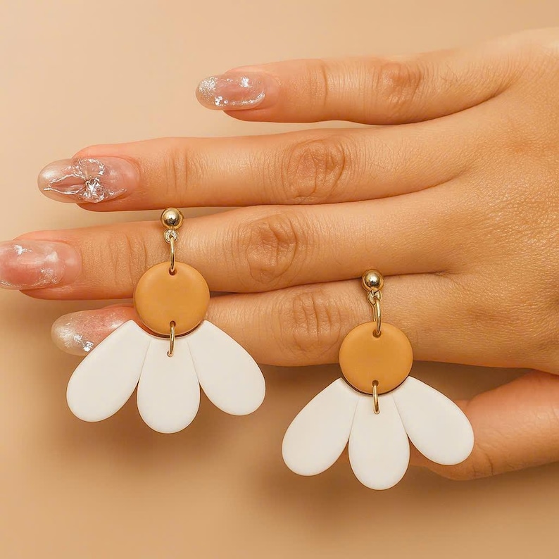 Puede incluir: Un par de pendientes dorados con una parte superior circular de color marr&oacute;n claro y una parte inferior blanca en forma de p&eacute;talo. Los pendientes est&aacute;n dise&ntilde;ados para parecerse a flores. Los pendientes se muestran sobre un fondo neutro.