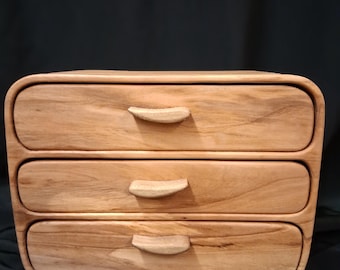 May include: A three-drawer wooden chest with a natural wood grain finish. The drawers have curved fronts and small, curved wooden handles. The chest has rounded edges and a smooth, polished appearance, suitable for storing small items.