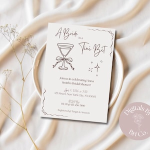 May include: A beige bridal shower invitation with the text "A Bride in a Tini Bit". The invitation features a martini glass illustration and event details. The invitation is on a white plate and a beige fabric background.