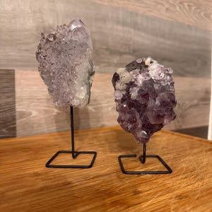 May include: Two amethyst crystal clusters on black metal stands. The crystals display shades of purple and white, with a textured, natural appearance. The stands have a square base and a thin, vertical support.