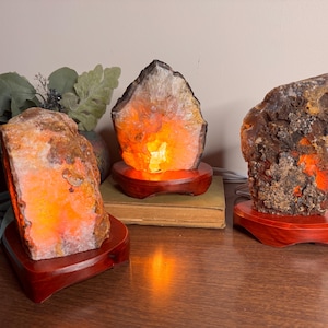 May include: Three illuminated agate lamps with warm orange and brown tones. Each lamp is a unique, natural stone formation, mounted on a dark red wooden base. The lamps are arranged on a wooden surface, creating a warm and inviting ambiance.