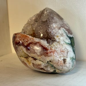 May include: A geode with a hollow center, showcasing a crystalline interior. The exterior features a mix of colors, including white, red, and green. The geode is displayed on a white surface.