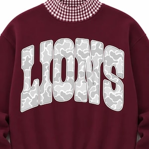 May include: A maroon long-sleeve sweatshirt featuring a high mock neck collar, cuffs, and a bottom hem in a white and maroon gingham pattern. The center of the chest displays the word LIONS in large, arched, white and gray camouflage letters.