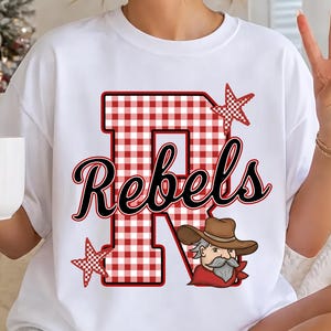 May include: A white short-sleeve t-shirt featuring a large red and white gingham letter R. The word Rebels is printed in black script across the letter. The design includes two red and white gingham stars and a cartoon cowboy with a brown hat.