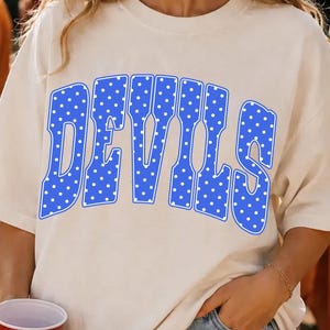 Puede incluir: Una camiseta de manga corta de color crema con la palabra DEVILS impresa en una fuente azul grande y arqueada. Las letras azules están rellenas con un patrón repetitivo de pequeños puntos blancos y delineadas con un borde azul fino.