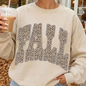 May include: A beige long-sleeve crewneck sweatshirt featuring the word FALL printed in large, bold, block letters with a brown and black leopard print pattern. The garment has a ribbed neckline and cuffs, and is worn with light blue denim shorts.