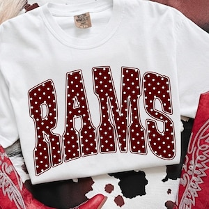 Puede incluir: Una camiseta blanca de manga corta con la palabra RAMS en letras grandes de estilo bloque color granate con un patrón de lunares blancos. La camiseta tiene cuello redondo y una etiqueta de marca en el cuello interior. El diseño está centrado.