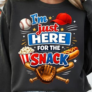May include: Dark grey sweatshirt featuring a baseball-themed graphic. The design includes a baseball, a red cap, popcorn, a hot dog, baseball bats, and a glove. Text reads "I'm just HERE for the SNACK."