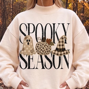 May include: A cream-coloured mock neck sweatshirt featuring the black text SPOOKY SEASON. The design includes a beige polka-dot ghost, a leopard-print pumpkin with brown leaves, and a black and white buffalo-check witch hat with a buckle.