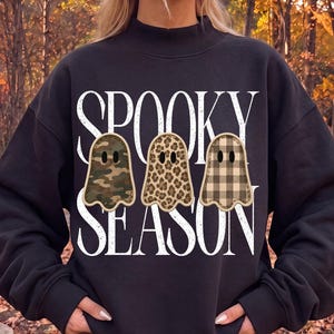 May include: A black long-sleeve sweatshirt featuring a mock neck and white text that reads SPOOKY SEASON. Three ghost-shaped graphics are centered on the chest, each with a different pattern: green camouflage, brown leopard print, and tan plaid.