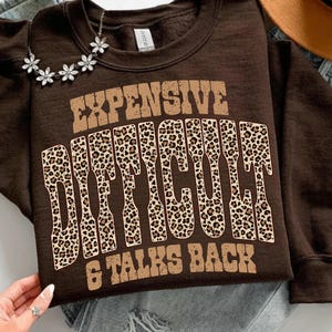 Puede incluir: Una sudadera marrón con el texto "EXPENSIVE DIFFICULT & TALKS BACK" en una fuente retro. La palabra "DIFFICULT" está rellena con un estampado de leopardo. Un collar plateado con colgantes en forma de flor está sobre la sudadera.