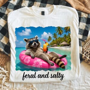 May include: A white t-shirt featuring a graphic of a raccoon wearing black sunglasses, floating on a pink hibiscus-patterned inner tube in turquoise ocean water, holding a tropical drink with a tiny umbrella. The text reads feral and salty.