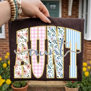 May include: A dark brown t-shirt featuring the word AUNT in large block letters with a gold outline. Each letter has a unique pattern: pink floral, pink gingham, blue floral, and blue stripes. The word aunt is written in cursive script.