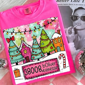 May include: A bright pink Comfort Colors t-shirt featuring a colorful Christmas design. The graphic includes decorated Christmas trees, houses, and the text "38008 bOliver, tenesse". A festive and cheerful holiday-themed apparel item.