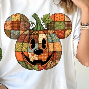 May include: White t-shirt featuring a colorful patchwork pumpkin design with Mickey Mouse ears. The pumpkin has a smiling face and is made up of various fall-themed fabrics in shades of orange, brown, and green. The shirt is a casual, oversized fit.