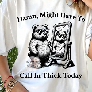 Op de afbeelding: Wit t-shirt met een zwart-witte illustratie van een beer met een zonnebril, kijkend in een spiegel. De tekst op het shirt luidt "Damn, Might Have To Call In Thick Today". Het t-shirt heeft een losse pasvorm.