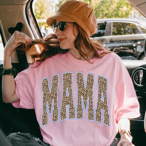 May include: A light pink short-sleeved t-shirt featuring the word MAMA printed in a large, bold, collegiate-style font. The letters have a brown and black leopard print pattern with a thin blue outline. The shirt is worn with a tan corduroy cap.