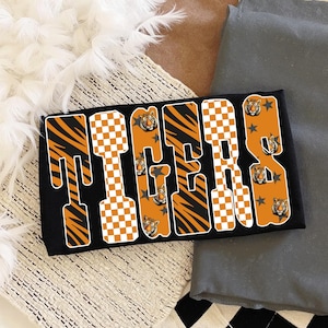 May include: Black rectangular design with the word "TIGERS" in orange and white checkered letters. The letters have tiger stripe accents and tiger head graphics. Gray stars and a tiger head are also included.