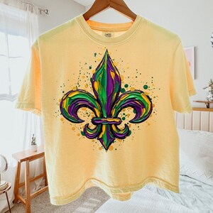 May include: Pale yellow short-sleeved t-shirt with a fleur-de-lis design in green, purple, and yellow. The graphic has textured brushstrokes and paint splatters. The t-shirt is hanging on a wooden hanger.