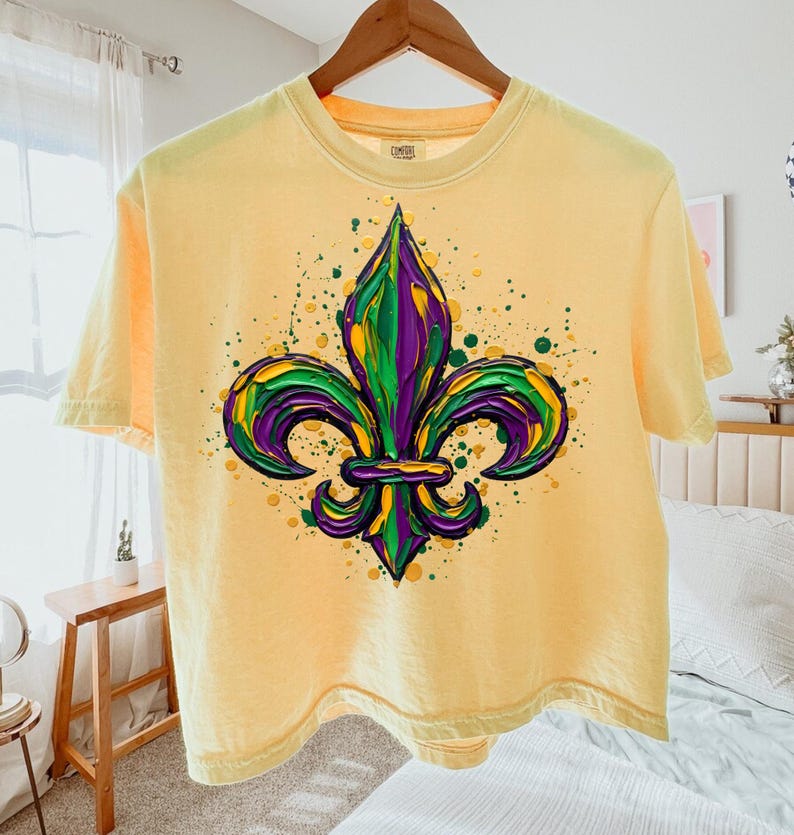 May include: Pale yellow short-sleeved t-shirt with a fleur-de-lis design in green, purple, and yellow. The graphic has textured brushstrokes and paint splatters. The t-shirt is hanging on a wooden hanger.