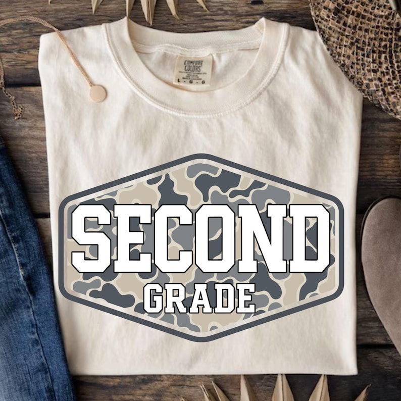 Pu&ograve; includere: Una t-shirt a maniche corte color crema con una grafica esagonale centrale caratterizzata da un motivo mimetico grigio, bianco e beige. Il testo "SECOND GRADE" &egrave; stampato in grassetto bianco con un contorno grigio scuro al centro.