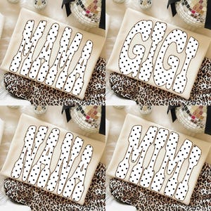 Può includere: Quattro magliette color crema con scritte a pois neri. Due magliette dicono "MAMA" e due dicono "GIGI". Le magliette sono su tessuto leopardato con palle da discoteca.