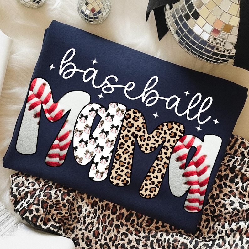 May include: Navy blue sweatshirt with the words "baseball mama" in white script. The word "Mama" is designed with baseball stitching, leopard print, and baseballs with bows. A fun, sporty design.