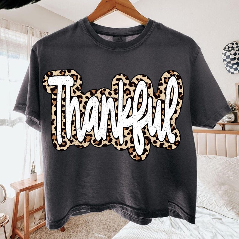Peut inclure: T-shirt gris fonc&eacute; avec le mot "Thankful" en lettres blanches. Le mot est soulign&eacute; d'un motif l&eacute;opard. Le t-shirt est suspendu &agrave; un cintre en bois.