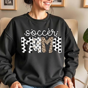May include: Black sweatshirt with the words "soccer mama" in white script and various patterns. The letters are decorated with soccer balls, dog faces, and leopard print. The sweatshirt has a crew neck and long sleeves.