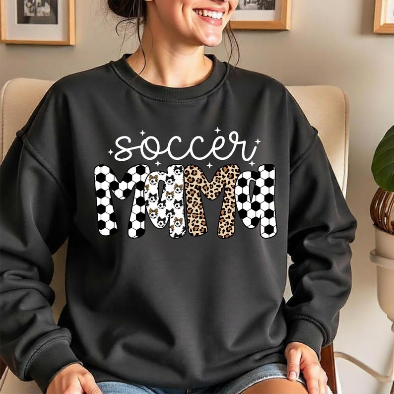 May include: Black sweatshirt with the words "soccer mama" in white script and various patterns. The letters are decorated with soccer balls, dog faces, and leopard print. The sweatshirt has a crew neck and long sleeves.