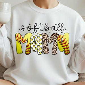 May include: White sweatshirt with the words "softball Mama" in black script and bold letters. The word "Mama" is designed with a softball, leopard print, and softball bow pattern. The sweatshirt is a casual style.