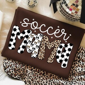 May include: A brown t-shirt with the words "Soccer Mama" in white script. The word "Mama" is designed with soccer ball patterns, leopard print, and soccer ball graphics. The shirt is folded and placed on a white surface with a leopard print fabric.