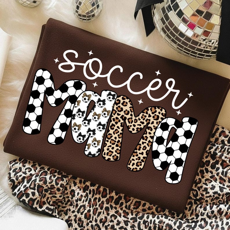 May include: A brown t-shirt with the words "Soccer Mama" in white script. The word "Mama" is designed with soccer ball patterns, leopard print, and soccer ball graphics. The shirt is folded and placed on a white surface with a leopard print fabric.