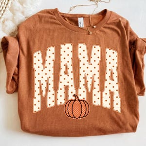 May include: A rust-colored short-sleeve t-shirt featuring the word MAMA in large, cream-colored block letters with brown polka dots. A small, orange pumpkin graphic with white polka dots is positioned below the text. The shirt has a crew neckline.