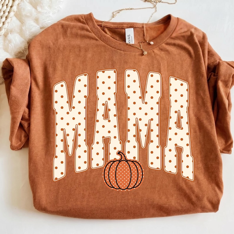 May include: A rust-colored short-sleeve t-shirt featuring the word MAMA in large, cream-colored block letters with brown polka dots. A small, orange pumpkin graphic with white polka dots is positioned below the text. The shirt has a crew neckline.