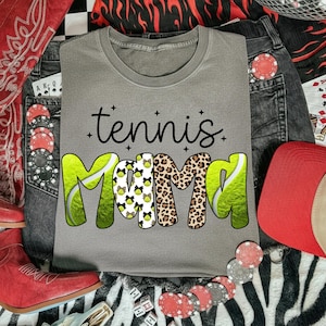 K&ouml;nnte beinhalten: Ein graues T-Shirt mit den Worten "tennis mama" in schwarzer Schrift und fetten, farbenfrohen Buchstaben. Das Wort "MAMA" zeigt ein Tennisball-Design, Leopardenmuster und Schleifen. Das Shirt ist von Jeans, Pokerkarten und einem roten Cowboyhut umgeben.