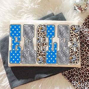May include: Cream-colored t-shirt with the word "EAGLES" in a bold, block letter design. The letters feature blue star patterns, a disco ball effect, and leopard print accents. Eagle head graphics are incorporated.