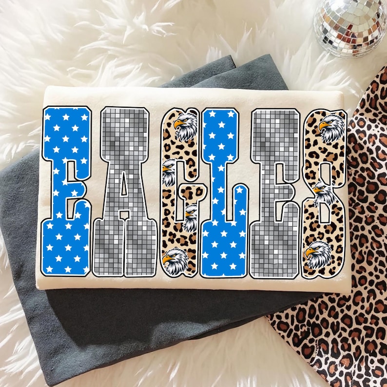 May include: Cream-colored t-shirt with the word "EAGLES" in a bold, block letter design. The letters feature blue star patterns, a disco ball effect, and leopard print accents. Eagle head graphics are incorporated.