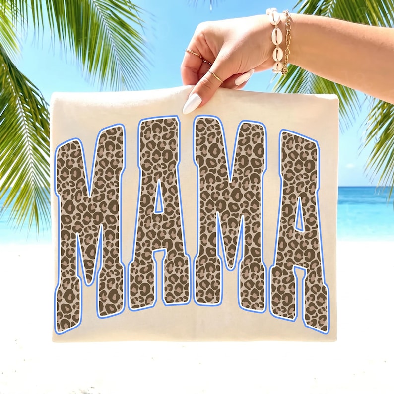 May include: A cream-colored t-shirt featuring the word MAMA in large, block-style letters. The letters have a brown and black leopard print pattern and are outlined in a light blue border. The design is centered on the front of the garment.
